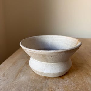 May include: A small, hand-thrown ceramic bowl with a wide, flared rim and a rounded, tapered base. The bowl features a speckled, off-white matte glaze with subtle brown earth-toned variations along the rim and the lower section of the body.