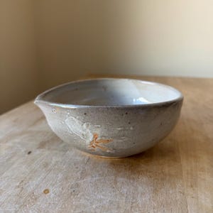 May include: A small, round ceramic bowl with a light gray, speckled glaze and a distinct pouring spout. The exterior features a subtle, embossed orange leaf-like pattern. The interior is smooth and off-white, with a curved rim and a sturdy base.