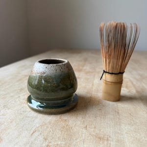 May include: A ceramic matcha whisk holder with a speckled cream top and a dark green glazed base, positioned next to a light brown bamboo matcha whisk featuring thin, flexible tines and a black thread binding around the central handle section.