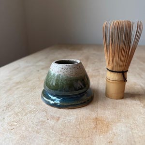 May include: A ceramic matcha whisk holder with a speckled cream top that transitions into a deep green and blue base, positioned next to a light-colored bamboo matcha whisk with thin, flexible tines tied with a black thread at the center.