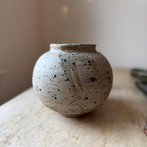 May include: A round ceramic vase with a textured, speckled beige surface featuring dark brown and black flecks. A smooth, glossy light green glaze drips from the rim down the side of the vessel, creating a contrast against the matte finish.