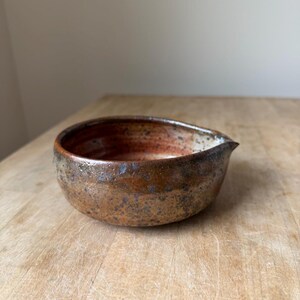 May include: A small, round ceramic bowl with a pour spout, featuring a mottled brown and rust-colored glaze with dark speckles. The interior transitions from a deep reddish-brown at the base to a lighter, cream-colored rim near the spout.