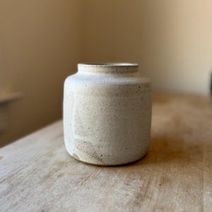 May include: A small, cylindrical ceramic vase with a matte, off-white finish and visible dark speckles throughout the surface. The vase features a short, slightly flared neck and a flat base, showcasing a textured, earthy appearance.