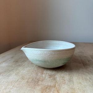 May include: A ceramic pouring bowl with a rounded shape and a small spout. The bowl features a speckled off-white glaze on the upper portion that transitions into a soft, muted green hue toward the base, creating a subtle gradient effect.