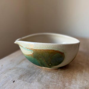 May include: A ceramic pouring bowl with a cream-colored crackle glaze finish. The exterior features a prominent, organic-shaped patch of blended green and brown earth-toned pigments. The bowl has a distinct, small spout on one side for pouring.