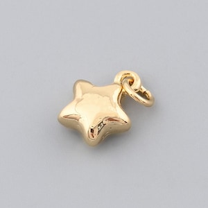 Puede incluir: Un pequeño dije de estrella dorada con un lazo para sujetarlo a un collar o pulsera. La estrella de cinco puntas tiene un diseño redondeado y tridimensional. El dije está sobre un fondo gris.