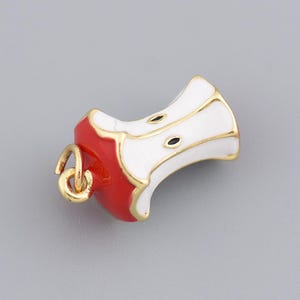 May include: A gold-coloured charm shaped like an apple core. The charm has a red base, white sides, and gold trim. Two small black dots represent seeds. A small gold loop is attached for hanging.