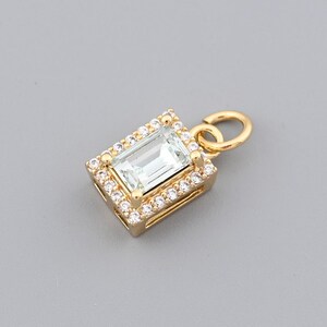 May include: A gold pendant with a rectangular, light blue gemstone at the center, surrounded by a halo of small, clear gemstones. The pendant has a small loop at the top for a chain.