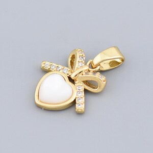 May include: A gold pendant with a white heart-shaped centre. The pendant has a bow design decorated with small, clear crystals. The bail is also gold. This pendant is likely for a necklace.