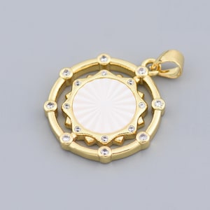 Puede incluir: Colgante dorado con un centro blanco texturizado y un marco circundante adornado con pequeñas piedras preciosas transparentes. El colgante tiene un diseño circular con un lazo para una cadena.