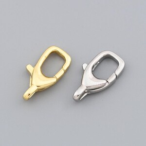 May include: Two metal lobster clasps, one gold-toned and one silver-toned, are displayed against a gray background. These clasps are commonly used in jewelry making for securing necklaces, bracelets, and other accessories.