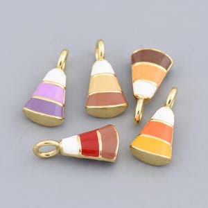 Può includere: Cinque ciondoli smaltati a forma di tubetti di vernice. Ogni ciondolo ha un'ansa color oro e presenta una diversa combinazione di colori: viola, beige, marrone, rosso e arancione. I ciondoli sono su sfondo grigio.