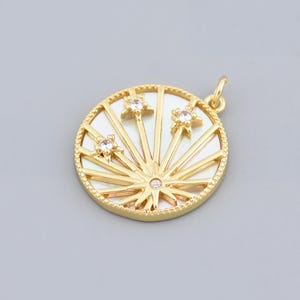 Puede incluir: Colgante dorado con diseño circular, fondo blanco y líneas radiantes. El colgante tiene tres adornos en forma de estrella con gemas transparentes y una pequeña gema en el centro. El colgante tiene un borde texturizado y un pequeño lazo para una cadena.