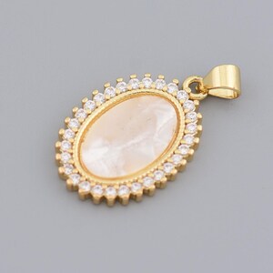 May include: Gold-toned pendant with an oval, pearlescent center stone. The stone is surrounded by a halo of small, clear crystals. The pendant has a simple bail for attaching to a chain. The jewelry is set against a gray background.