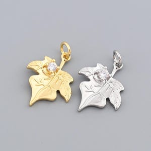May include: Two leaf-shaped pendants, one gold-toned and one silver-toned, each with a small, clear gemstone at the center. The pendants have a bail at the top for attaching to a chain or cord. The background is a solid gray.