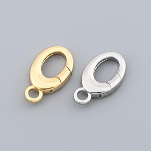 Puede incluir: Dos cierres de langosta ovalados, uno dorado y otro plateado, se muestran sobre un fondo gris. Cada cierre tiene un pequeño lazo en la parte inferior para sujetarlo a joyas u otros artículos. Los cierres están cerrados, mostrando su mecanismo de cierre seguro.