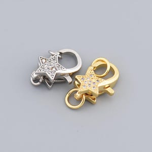 Puede incluir: Dos cierres en forma de estrella, uno plateado y otro dorado, se muestran sobre un fondo gris. Las estrellas están adornadas con pequeñas piedras brillantes. Cada cierre tiene un anillo y un gancho.