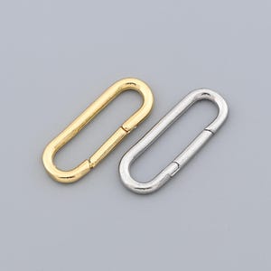 May include: Two metal oval-shaped clasps. One is gold-colored, and the other is silver-colored. The clasps are positioned on a gray background. These clasps are likely used for attaching straps or closures on bags, accessories, or clothing.