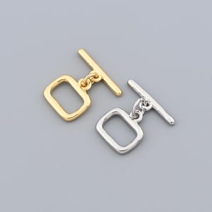 Op de afbeelding: Twee tuimelsluitingen, één goudkleurig en één zilverkleurig, tegen een grijze achtergrond. Elke sluiting heeft een rechthoekige lus en een staaf, geschikt voor het maken van sieraden of knutselprojecten. Ideaal voor armbanden en kettingen.