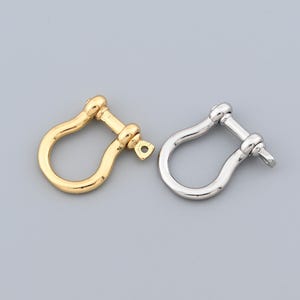 May include: Two metal shackle clasps, one gold-toned and one silver-toned, are displayed against a gray background. Each clasp has a U-shaped body with a screw pin closure. The gold clasp has a small loop.