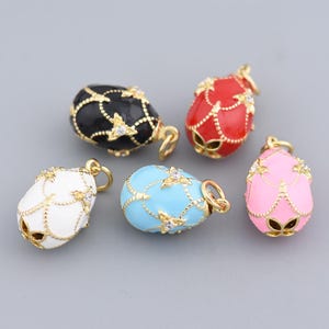 Könnte beinhalten: Fünf eiförmige Anhänger in Schwarz, Rot, Weiß, Blau und Rosa. Jeder Anhänger hat eine goldfarbene Umrandung und ist mit einem goldfarbenen Kettendesign und kleinen, klaren Kristallen verziert.