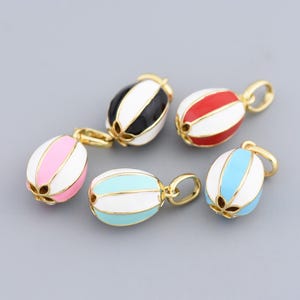 Könnte beinhalten: Fünf bunte Emaille-Anhänger mit goldfarbenen Schlaufen. Die ovalen Anhänger haben vertikale Streifen in Schwarz, Rot, Rosa, Hellblau und Weiß. Der Hintergrund ist einfarbig grau.