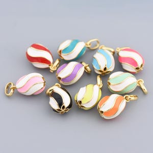 Könnte beinhalten: Eine Sammlung bunter Emaille-Anhänger mit goldfarbenen Fassungen. Jeder Anhänger ist eiförmig mit weißen und gestreiften Designs in verschiedenen Farben, darunter Rot, Rosa, Lila, Blau, Grün, Schwarz, Gelb und Orange.