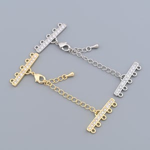 May include: Three multi-strand necklace clasps in gold and silver tones. Each clasp features a bar adorned with small, clear crystals and multiple loops for attaching strands. The clasps include a lobster claw closure and an extender chain.