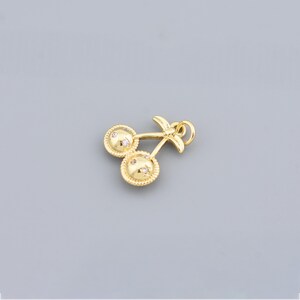 May include: A gold-coloured cherry charm featuring two round cherry halves, each with small, clear stones. The charm has a stem and a loop for attaching to a necklace or bracelet. The background is a solid grey.