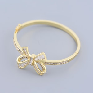 Puede incluir: Un brazalete de oro con un diseño de lazo de cristal transparente. El brazalete está hecho de metal y tiene un acabado pulido.