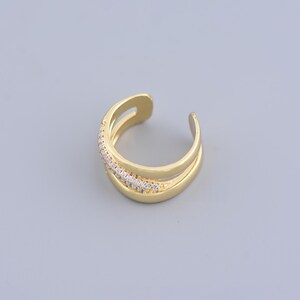 May include: A gold-colored ear cuff with a layered design. One layer is adorned with small, clear gemstones. The jewelry piece has a curved shape and is designed to be worn on the ear.