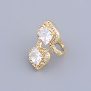 May include: A gold ring with two large, clear, square-shaped cubic zirconia stones. The ring is designed with a double band and features smaller cubic zirconia stones around the edges.