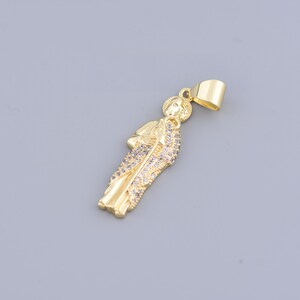 May include: A gold-colored pendant in the shape of a religious figure. The pendant features a detailed design with sparkling embellishments on the robe. The bail is a simple, rectangular loop for attaching to a chain. The pendant is set against a gray background.