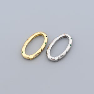 May include: Two oval-shaped clasps, one gold-toned and one silver-toned, are displayed against a gray background. Both clasps are adorned with small, colorful gemstones. The gold clasp has a polished finish, while the silver clasp has a metallic sheen. These clasps are likely used for jewelry making.
