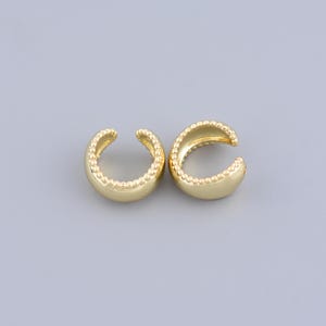 May include: A pair of gold ear cuffs with a beaded design. The cuffs are made of metal and have a smooth, polished finish.