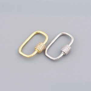 May include: Two oval-shaped carabiner clasps, one gold-toned and one silver-toned, are displayed against a gray background. Each clasp features a cylindrical screw-lock mechanism encrusted with small, sparkling stones, adding a touch of elegance to the design.