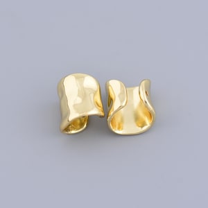 May include: Two gold-coloured ear cuffs with an abstract shape. The ear cuffs have a smooth, reflective surface and a curved design. The jewellery is displayed on a grey background.