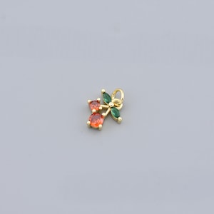 May include: A gold-coloured charm with a cherry design. It features two round, orange-red gemstones for the cherries and four green, marquise-cut gemstones as leaves. A small loop at the top allows attachment to a chain.