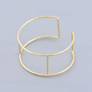 Può includere: Un braccialetto rigido in metallo dorato con un design geometrico. Il braccialetto è aperto e ha uno stile semplice e minimalista.