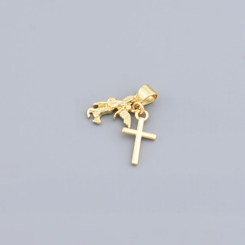 May include: A gold-colored pendant featuring a small winged horse and a cross charm. The pendant has a loop for a chain and is set against a gray background. The cross is a simple, classic design.