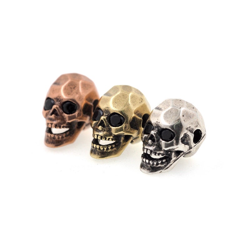 May include: Three miniature skull-shaped beads, each with a black gemstone eye. The skulls are made of different metals: copper, brass, and silver.