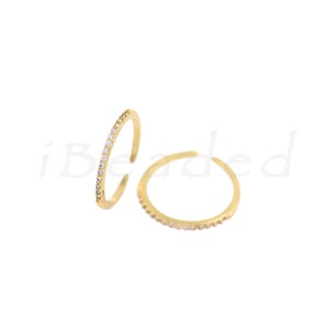May include: Two gold-colored adjustable rings. One ring is in focus, showing a row of small, clear gemstones. The other ring is slightly out of focus. Both rings are on a white background. The rings are delicate and minimalist.