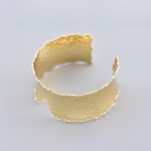 Op de afbeelding: Een goudkleurige manchetarmband met een getextureerd oppervlak en een golvende, onregelmatige rand. Het open ontwerp maakt het gemakkelijk te dragen. Dit sieraad is een opvallend accessoire.