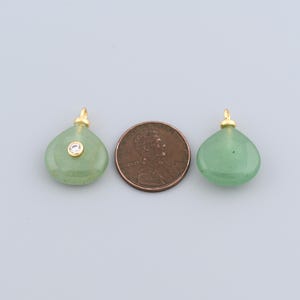 May include: Two green teardrop-shaped pendants with gold-coloured bails. One pendant has a small, clear gemstone. A penny is placed between the pendants for size comparison. The pendants are likely for jewellery making.