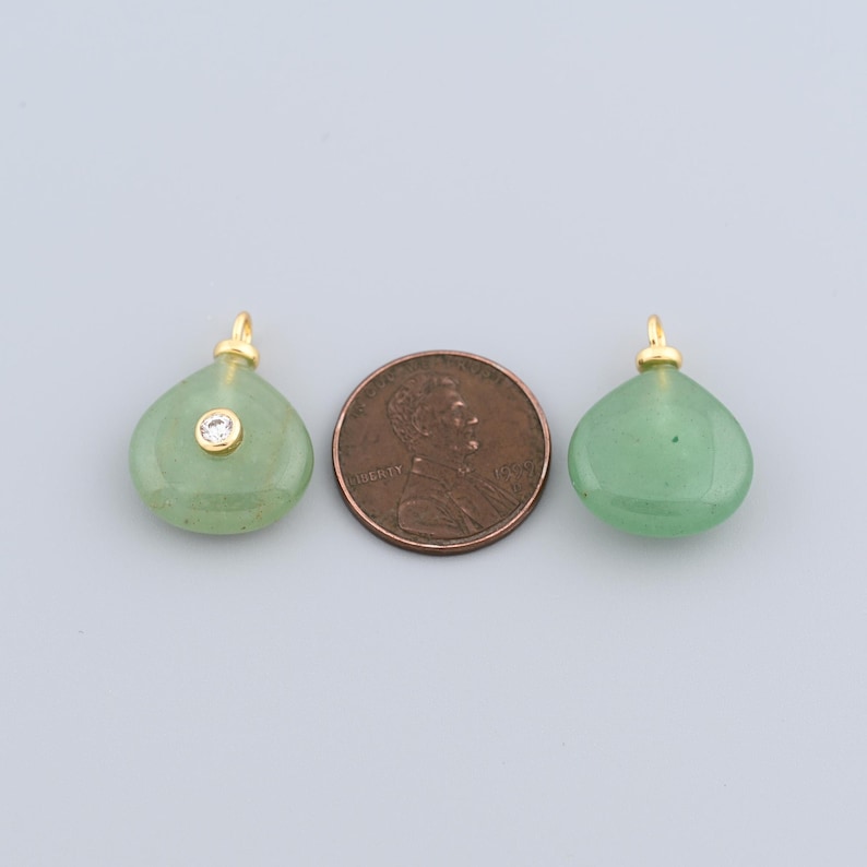 May include: Two green teardrop-shaped pendants with gold-coloured bails. One pendant has a small, clear gemstone. A penny is placed between the pendants for size comparison. The pendants are likely for jewellery making.