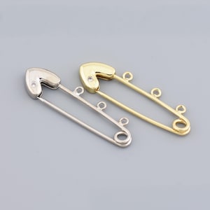 May include: Two safety pins with heart-shaped heads. One is silver-colored, the other is gold-colored. Both have a small, clear gem set in the heart. The pins have a decorative loop at the end and a small loop in the middle.