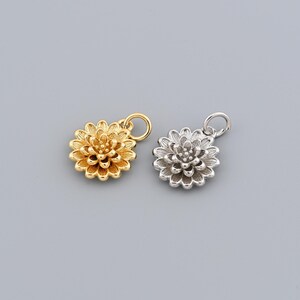 Può includere: Due ciondoli a forma di fiore, uno dorato e uno argentato. Ogni ciondolo presenta un design floreale dettagliato e un piccolo anello nella parte superiore per il fissaggio a una catena o un braccialetto. Lo sfondo è grigio.