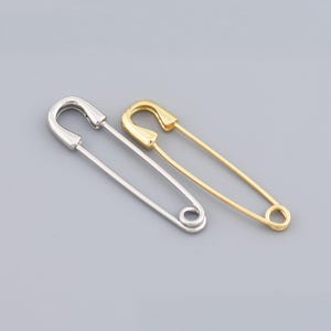 Può includere: Due spille di sicurezza, una argentata e una dorata, sono mostrate su uno sfondo grigio. Le spille hanno un design identico, con una parte superiore curva e una base arrotolata. La spilla dorata ha una finitura lucida, mentre quella argentata è opaca.