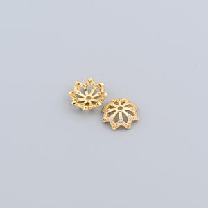 Peut inclure: Deux capuchons de perles décoratifs dorés avec un motif floral. Chaque capuchon présente un motif ajouré et de petites pierres précieuses transparentes. Les capuchons sont présentés sur un fond gris clair, mettant en valeur leurs détails complexes.