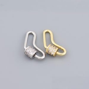 Può includere: Due chiusure decorative, una argentata e una dorata, ciascuna con un design a gancio curvo. La chiusura argentata presenta un cilindro incastonato con cristalli trasparenti. La chiusura dorata ha un cilindro simile con cristalli trasparenti. Su sfondo grigio.